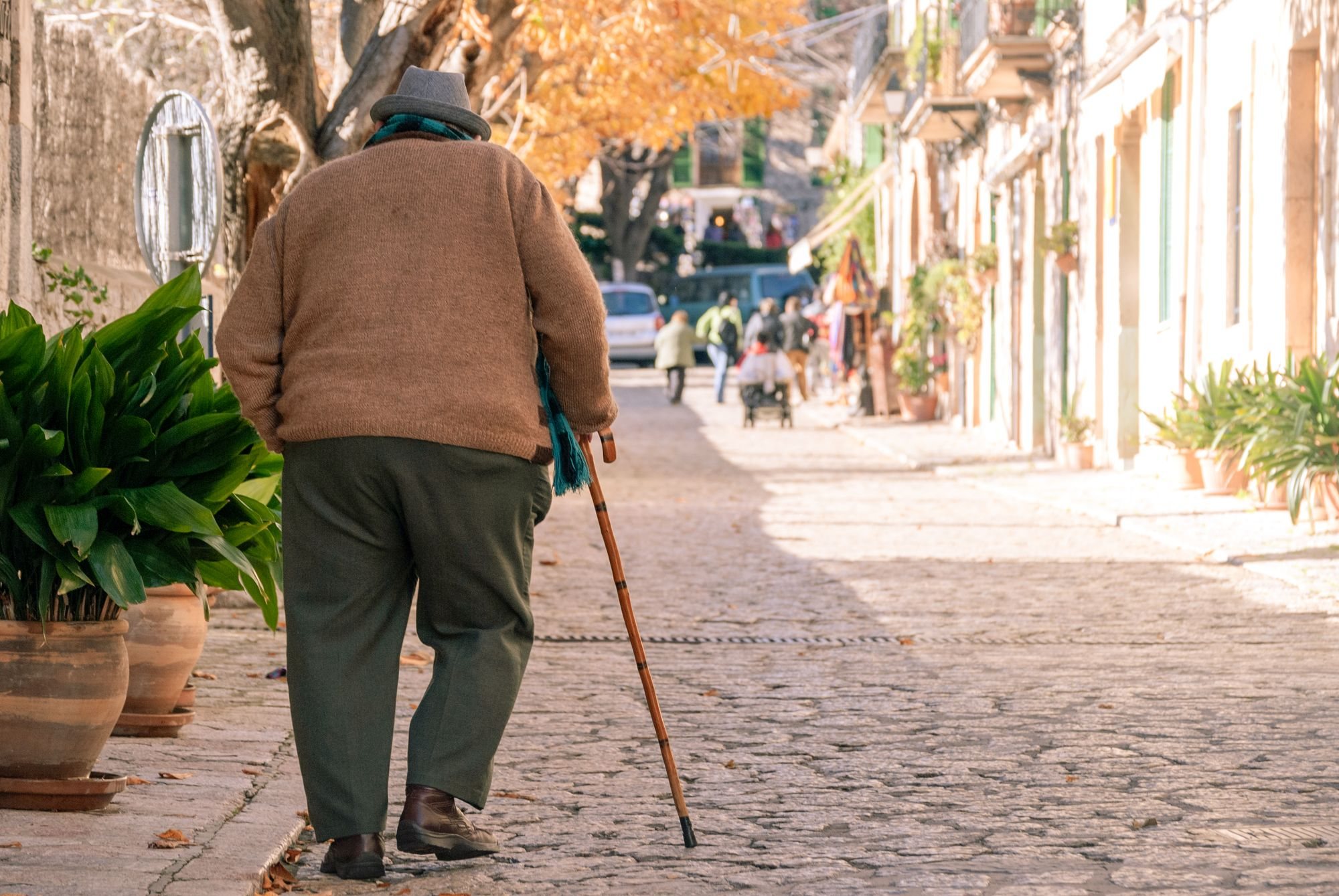 Senhor de costas caminhando com bengala por uma rua de pedras